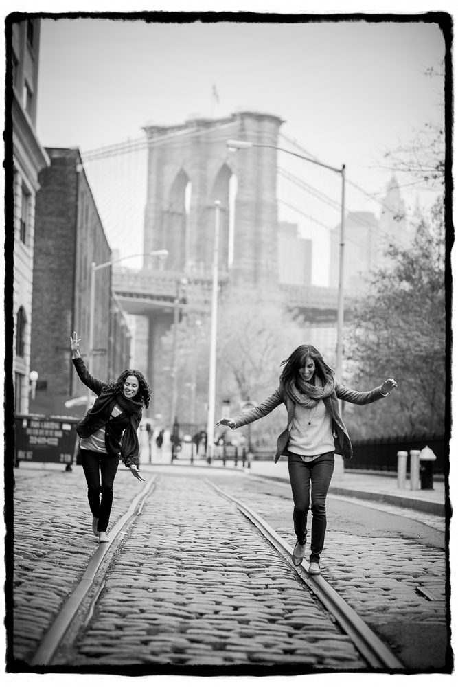 Engagement Portraits: Lara & Nicole on a cobblestone street with the Brooklyn Bridge behind them.